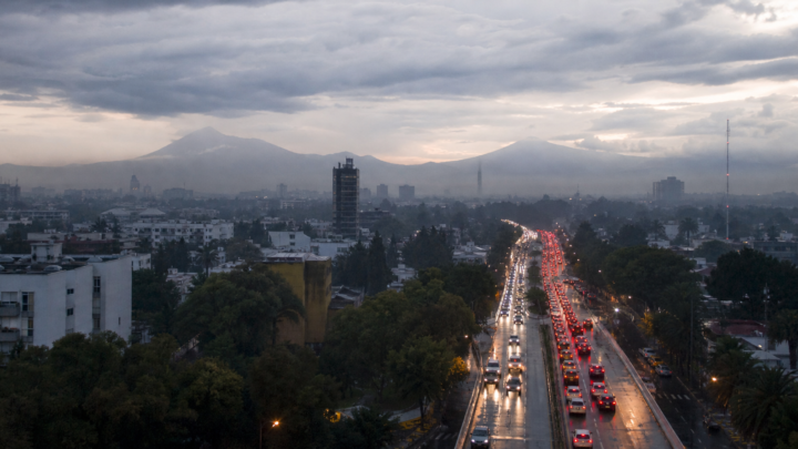 Tráfico vehicular en una avenida de la Ciudad de México al amanecer, con pavimento mojado y cielo nublado.