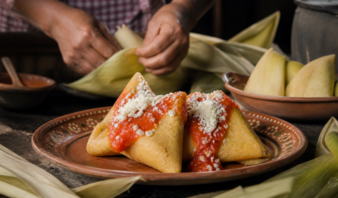 Corundas triangulares de maíz servidas con salsa roja, queso y hojas de caña sobre una mesa de cocina tradicional.